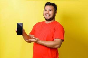 Fröhlicher dicker asiatischer Mann präsentiert Handy mit leerem Bildschirm isoliert auf gelbem Hintergrund foto