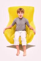 Porträt eines coolen jungen, stilvollen Jungen, der auf gelbem Stuhl auf hellem Hintergrund sitzt foto