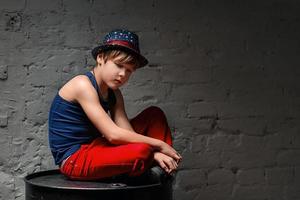 Porträt eines coolen jungen Hip-Hop-Jungen mit blauem Hut und roter Hose, der auf einem schwarzen Fass im Dachboden sitzt foto