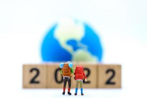2022 Neujahr und Reisekonzept. Nahaufnahme von zwei Miniaturfiguren des Reisenden mit Rucksack, der mit Stapel aus Holznummernblock und Mini-Weltkugel auf weißem Hintergrund steht. foto