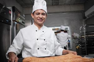 älterer asiatischer männlicher koch in weißer kochuniform und hut, der ein tablett mit frischem, leckerem brot mit einem lächeln zeigt, in die kamera schaut, glücklich mit seinen gebackenen lebensmitteln, professioneller job in der edelstahlküche. foto