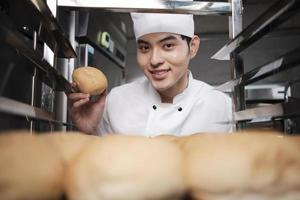 junger asiatischer männlicher koch in weißer kochuniform und hut, der mit einem lächeln ein tablett mit frischem leckerem brot zeigt, in die kamera schaut, glücklich mit seinen gebackenen lebensmitteln, professioneller job in der edelstahlküche. foto