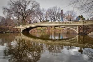 die bogenbrücke im central park, new york city tageslichtansicht mit reflexion im wasser, wolken, bäumen und manhattan skyline im hintergrund foto