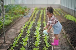 Kleines Afro-Kind-Bauernmädchen im Garten, das Pflanzen wässert und Gemüse erntet. foto