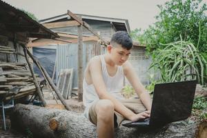 Armer Schuljunge, der online mit Notizbuch auf Baumstamm hinter einem alten Landhaus studiert. konzept online-bildung in ländlicher umgebung und arbeit von zu hause aus foto