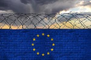 ziegelmauer mit stacheldraht in den farben der flagge der europäischen union, eu, gegen einen stürmischen himmel. Krise und hybrider Krieg, Grenzschließungen. foto
