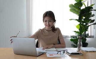 asiatische geschäftsfrau, die mit einem kollegenteam in einer videoanrufkonferenz spricht und eine notiz auf einem buch mit einem lächelnden gesicht schreibt. frau, die computer-laptop und kopfhörer für online-meetings verwendet. foto