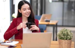 junge asiatische geschäftsfrau hat das vergnügen, die arbeit auf ihrem tablet und ihrem lieblingskaffee im büro zu beobachten. foto