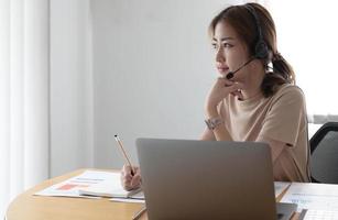 asiatische geschäftsfrau, die mit einem kollegenteam in einer videoanrufkonferenz spricht und eine notiz auf einem buch mit einem lächelnden gesicht schreibt. frau, die computer-laptop und kopfhörer für online-meetings verwendet. foto