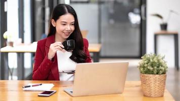 junge asiatische geschäftsfrau hat das vergnügen, die arbeit auf ihrem tablet und ihrem lieblingskaffee im büro zu beobachten. foto