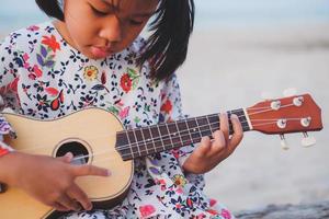 junges asiatisches mädchen, das ukulele am strand spielt. foto