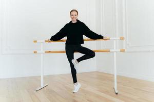 horizontale aufnahme einer flexiblen jungen frau lehnt sich an ballettstange, trägt schwarzes kostüm und weiße sportschuhe, lächelt positiv, posiert im tanzsaal. Ballettlehrerin demonstriert verschiedene Übungen foto