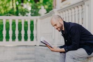 Menschen, Hobby und Lifestyle-Konzept. Positiver Glatzkopf liest mit Begeisterung ein Buch, posiert im Freien vor einem weißen Balkon, hat einen fröhlichen Ausdruck, genießt eine ruhige Atmosphäre. hübsche männliche studien mittleren alters foto