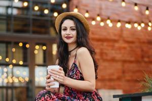 Außenaufnahme einer hübschen Frau mit dunklen langen Haaren, reiner, gesunder Haut, Strohhut und Kleid, die heißen Tee trinkt, während sie auf der Terrasse der gemütlichen Cafeteria sitzt. schöne Frau, die sich im Café ausruht foto