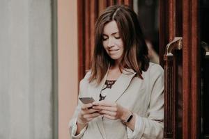 Zufriedene Geschäftsfrau hält ein modernes Handy in der Hand, gekleidet in eine weiße Jacke, durchsucht das Internet, ist mit WLAN verbunden und posiert im Freien. Unternehmerin nutzt Online-Banking, überweist Geld foto