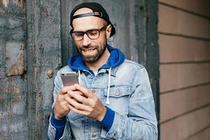 horizontales porträt eines gutaussehenden bärtigen, stilvollen mannes im jeansanorak mit großer brille, der ein smartphone mit kostenlosem internet hält, e-mails checkt, lustige videos ansieht, anwendungen aktualisiert foto