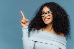 zufrieden lächelnde dunkelhäutige frau mit afro-frisur zeigt zeigefinger nach oben, demonstriert etwas auf leerraum, hat frohen ausdruck, trägt blauen pullover, steht drinnen. Menschen und Förderung foto