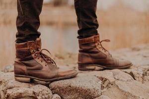 beschnittene aufnahme eines nicht erkennbaren mannes trägt alte schuhe, hat einen spaziergang im freien, unscharfer hintergrund. Stiefel auf Steinen. Modekonzept foto