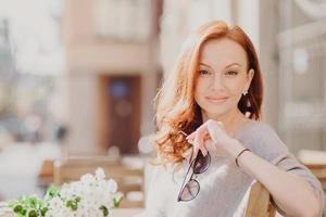 horizontale aufnahme einer attraktiven, erfreuten rothaarigen frau sitzt auf einer bank, genießt den sonnigen tag, hält eine sonnenbrille, atmet frische luft, hat make-up, posiert auf einem verschwommenen straßenhintergrund. Menschen und Lebensstil foto