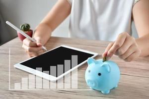 Eine Frau legt eine Münze in ein blaues Sparschwein auf einen Holztisch, um Geld zu sparen und das Haushaltseinkommen auf dem Tablet aufzuzeichnen. foto