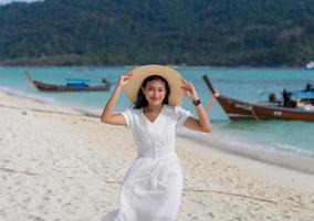 Porträt einer attraktiven asiatischen Frau in weißem Kleid und Hut mit breiter Krempe, die vor Glück am blauen Meeresstrand lächelt foto
