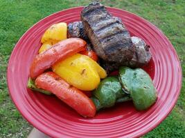 Hand, die einen roten Teller mit gegrilltem Steak und roten, gelben und grünen Paprika hält foto