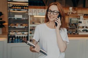 Fröhliche, schöne Geschäftsfrau, die draußen mit dem Handy spricht und mit Laptop in der Nähe des Cafés steht foto