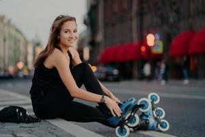 fit positive Frau zieht Rollerblades an sitzt auf Asphalt vor verschwommenem Straßenhintergrund hat einen positiven Ausdruck geht für Sport ein hat Fitnessaktivitäten im Freien während der Sommerzeit. Hobby-Konzept foto