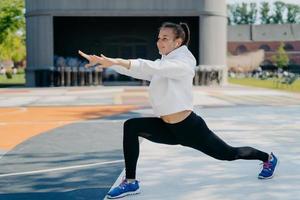 Außenaufnahme einer schlanken, sportlichen Frau, die an Freizeitaktivitäten beteiligt ist, streckt die Arme, Beine, blickt in die Ferne, trägt weiße Hoodie-Leggings und Sportschuhe, dehnt den Körper, bevor der Lauf zu einem gesunden Lebensstil führt foto