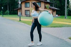 Ganzkörperaufnahme einer sportlich schlanken Frau mit großem, aufgeblasenem Fitball, in Aktivkleidung gekleidet, macht Aerobic-Übungen im Freien, hat einen athletischen Körper, der auf der Straße steht, mit Haus im Hintergrund. Menschen, Lebensstil foto