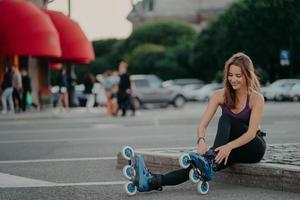 sportlicher Lifestyle und Hobby. Zufriedene dunkelhaarige Europäerin zieht Inline-Skates an und geht Inline-Skating-Posen vor verschwommenem Stadthintergrund, hält sich fit und verbringt ihre Freizeit aktiv. Außenaufnahme