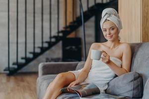 Das Foto einer entzückenden jungen Frau hat Make-up und gesunde Haut, hält eine Tasse Kaffee in der Hand, ist in ein Badetuch gewickelt, liest eine Zeitschrift und fühlt sich entspannt auf dem Sofa, verbringt Freizeit zu Hause. Trost empfinden