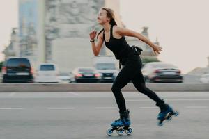 Junge, dunkelhaarige, schlanke, gesunde Frau hat Erholungszeit, genießt Rollblading auf Geschwindigkeit, bewegt sich schnell, hat einen fröhlichen Ausdruck, trägt ein schwarzes T-Shirt und Leggings. aktives ruhe- und sporttrainingskonzept foto