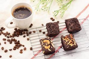 4 quadratische dunkle Brownies mit Schokoladensplittern, Mandeln und Nüssen auf einem Backgitter, 1 weiße Tasse Kaffee und Kaffeebohnen auf einem weißen Tuch. foto