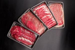 Gruppe von gemischtem rohem Rindfleisch. In Scheiben geschnittenes rohes Rindfleisch in einer Reihe auf einem schwarzen Kunststofftablett, das auf schwarzem Hintergrund isoliert ist. foto