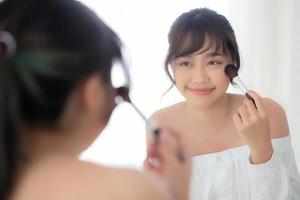 schönheitsporträt junge asiatische frau lächelt mit gesicht, das spiegel beim schminken mit bürstenwange im schlafzimmer anwendet, schön von mädchen, die blusher, hautpflege und kosmetisches konzept halten. foto