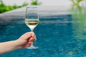 Hand, die ein Glas Weißwein mit Schwimmbadhintergrund hält. Urlaubs- und Sommergetränkekonzept. foto