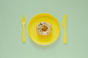 gebissener Donut auf gelbem Teller mit Messer und Gabel auf grünem Hintergrund. minimales kreatives ungesundes lebensmittelkonzept. foto