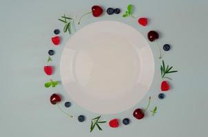 frische kirsche, blaubeere, himbeere, minze und rosmarinblatt auf draufsicht mit weißer platte und pastellblauem hintergrund für ein gesundes lebensmittelkonzept. foto