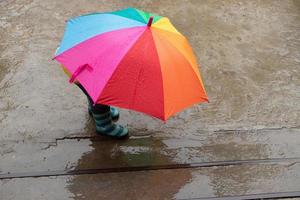 Ein 3-jähriges Mädchen versteckt sich im Regen unter einem farbigen Regenschirm foto
