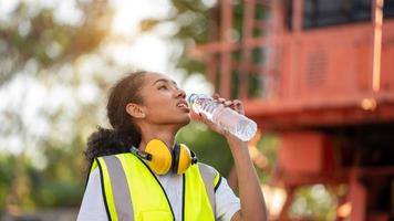 Abweichend trinkt eine afroamerikanische Vorarbeiterin oder Arbeiterin mit Cross-Eye-Sicherheit eine Flasche Wasser, nachdem sie die Arbeit beendet und sich auf dem alten Lastwagen am Frachtcontainerhafen entspannt hat foto