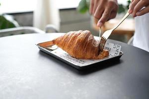 Nahaufnahme der Hand verwenden Gabel und Messer, um ein Stück Croissant zu schneiden, um es zum Frühstück auf einem Holztisch im Café zu essen. foto