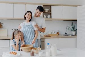 glückliche familie kocht zusammen in der küche. vater, mutter und tochter sind damit beschäftigt, köstliche mahlzeiten zu hause zuzubereiten. mann umarmt frau, die teig schlägt und zubereitet, kekse backt. essen, zusammensein foto