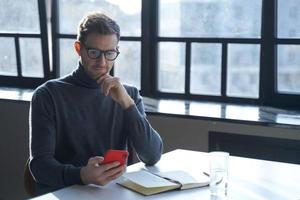 junger deutscher geschäftsmann, der internetnachrichten liest oder e-mails auf dem smartphone checkt, während er am schreibtisch sitzt foto