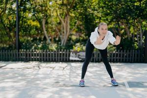 Außenaufnahme einer zufriedenen, gesunden, sportlichen Frau, die sich nach vorne lehnt, hält die Handflächen erhoben, macht Übungen in Open-Air-Zügen im Park, fühlt sich gut an, trägt Sweatshirt-Leggings und Turnschuhe, führt einen aktiven Lebensstil foto