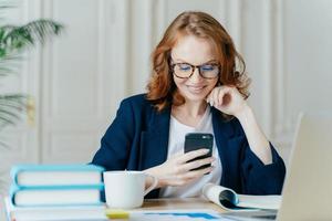 Foto einer hübschen Frau sitzt mit Smartphone-Gerät, gibt Feedback ein, arbeitet im Büro an einem aktuellen Laptop, konzentriert sich auf den Bildschirm des Gadgets, sitzt am Arbeitsplatz mit Büchern, Notizblock und heißem Getränk