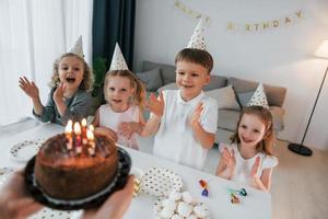 Frau, die Kuchen mit Kerzen hält. Geburtstag feiern. Gruppe von Kindern ist tagsüber zusammen zu Hause foto