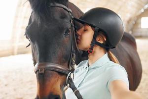 Schönes Selfie machen. eine junge frau in jockeykleidung bereitet sich auf einen ritt mit einem pferd auf einem stall vor foto