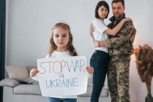 Mädchen mit Banner mit Stop-War-Text darauf. Soldat in Uniform ist mit Frau und Tochter zu Hause foto