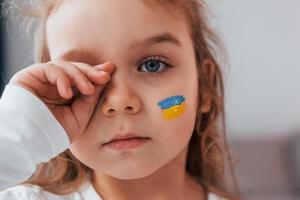 Unschuldiges Kind weint. Porträt eines kleinen Mädchens mit ukrainischer Flagge im Gesicht foto